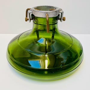 May include: A green glass Christmas tree stand with a metal collar and adjustable brackets. The stand has a rounded base and a wide opening for the tree trunk. The metal collar is silver-colored.