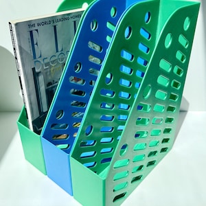 May include: A three-section magazine holder in green and blue plastic. The holder has a slotted design and holds a magazine with the text "ELLE DECOR". The magazine holder is designed to organize magazines and periodicals.