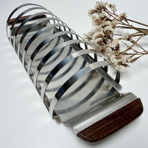 May include: A stainless steel toast rack with a rectangular base and curved metal bars. The base and handle have dark wood accents. Dried flowers are in the background. The rack is approximately 20 cm long.