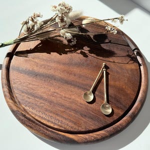 Puede incluir: Una tabla redonda de madera marrón oscuro con borde tallado. Dos pequeñas cucharas doradas descansan sobre la tabla. Una ramita de flores secas se coloca en el borde, agregando un acento natural.