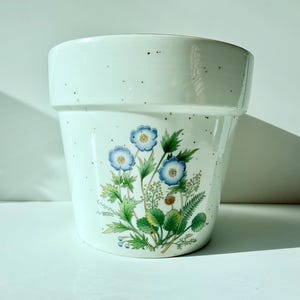 May include: A white ceramic flower pot with a speckled finish and a wide rim. The front features a detailed botanical illustration of blue flowers with green leaves, stems, and small buds, rendered in a realistic style on the smooth surface.