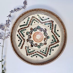 May include: A round ceramic plate with a cream-colored base featuring a central terracotta circle surrounded by dark brown and teal painted patterns. The rim has a textured, scalloped edge in brown and cream, with a sprig of purple flowers nearby.