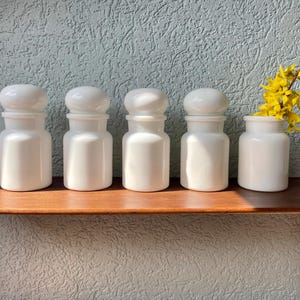 May include: Five white ceramic apothecary jars with rounded lids and a small vase holding yellow flowers are displayed on a wooden shelf. The jars are arranged in a row against a textured, light blue wall, creating a clean and organized aesthetic.