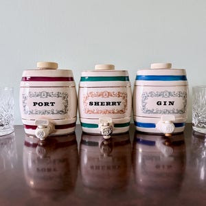May include: Three ceramic barrel-shaped liquor dispensers labeled "Port", "Sherry", and "Gin". Each dispenser has a spigot and is decorated with colored bands and text. Two crystal glasses are on either side.