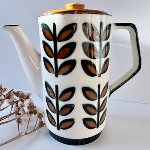 May include: A tall, white ceramic coffee pot with a brown lid and handle. The pot features a repeating pattern of brown leaf-like shapes outlined in black. The spout and handle are white, and the base is black.