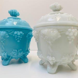 Könnte beinhalten: Zwei antike Glasschalen mit Deckeln. Eine ist aus weißem Milchglas und die andere ist hellblau. Beide haben ein Traubenmuster und drei Füße.