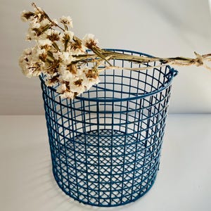 Op de afbeelding: Een ronde, donkerblauwe draadmand met een rasterpatroon, met een bos gedroogde, crèmekleurige bloemen. De mand is cilindrisch en staat op een wit oppervlak.