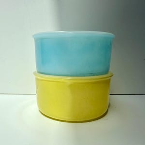 May include: Two round glass bowls stacked vertically. The top bowl is a translucent light blue, while the bottom bowl is a solid, opaque yellow. Both bowls feature smooth, rounded rims and straight sides, creating a simple, functional design.
