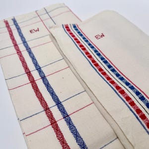 May include: Three cream-coloured kitchen towels with red and blue decorative stripes and diamond patterns. Each towel has the embroidered initials "EW". The towels are folded to display the design details.