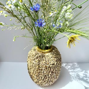 May include: A round, textured ceramic vase with a rough, sandy-colored exterior and a smooth, yellow-rimmed opening. The vase is filled with a vibrant bouquet of wildflowers, including white daisies, bright blue cornflowers, and green wheat stalks.