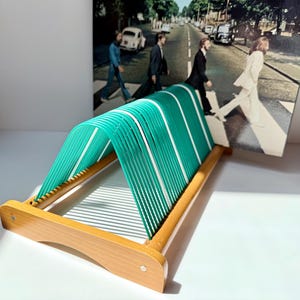 May include: A wooden record holder with a green and white striped design. The holder has a rectangular wooden frame and green and white plastic strips. The background features a framed image of the Beatles crossing Abbey Road.