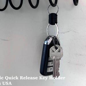 May include: A black magnetic quick-release key holder with a car key and a house key attached. The key holder is hanging from a black, curved metal bar. The text "Magnetic Quick Release Key Holder Made in USA" is at the bottom.