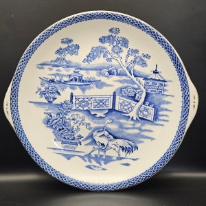 Könnte beinhalten: Ein runder, weißer Servierteller mit blauem und weißem Chinoiserie-Design. Das Muster zeigt eine Landschaft mit einem Boot, Bäumen und Vögeln. Der Rand hat eine gewellte Bordüre. Der Teller hat zwei kleine Griffe mit goldenen Akzenten.