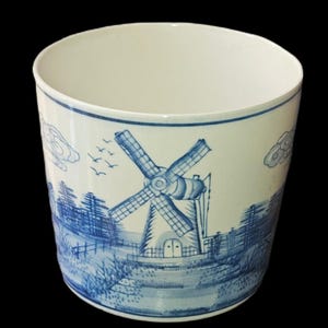 May include: A cylindrical ceramic planter with a white base and blue hand-painted illustrations. The design features a prominent windmill, stylised trees, clouds, and birds in flight. The interior of the vessel is plain white with a smooth finish.
