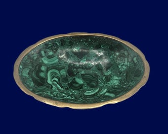 May include: An oval-shaped bowl featuring a deep green malachite stone interior with intricate swirling patterns and a scalloped brass metal rim. The bowl has a polished finish that highlights the natural banding and rich color variations of the stone.