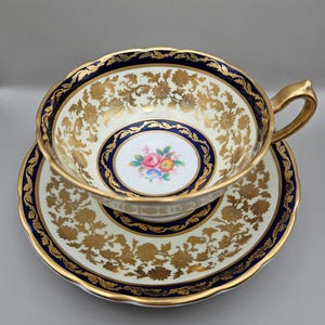 Puede incluir: Un juego de taza y platillo de té vintage con un patrón floral dorado y azul. La taza y el platillo tienen un fondo blanco con un diseño floral dorado y un borde azul. El centro del platillo tiene un motivo floral.