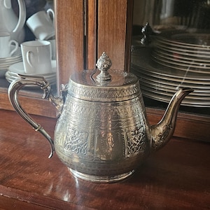 May include: A silver-toned metal teapot featuring an ornate lid with a decorative finial. The body is adorned with intricate embossed panels depicting figures and geometric patterns, complemented by a curved handle and a tapered spout.