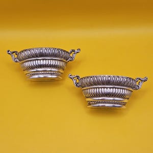 May include: Two silver bowls with decorative handles and fluted detailing. The bowls have a decorative design around the rim and base. The background is a solid yellow colour.