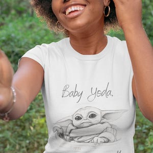 May include: White t-shirt featuring a sketch of Baby Yoda with the text "Baby Yoda, my name is not!" The shirt is worn by a person with dark curly hair and gold hoop earrings. The background is blurred green.