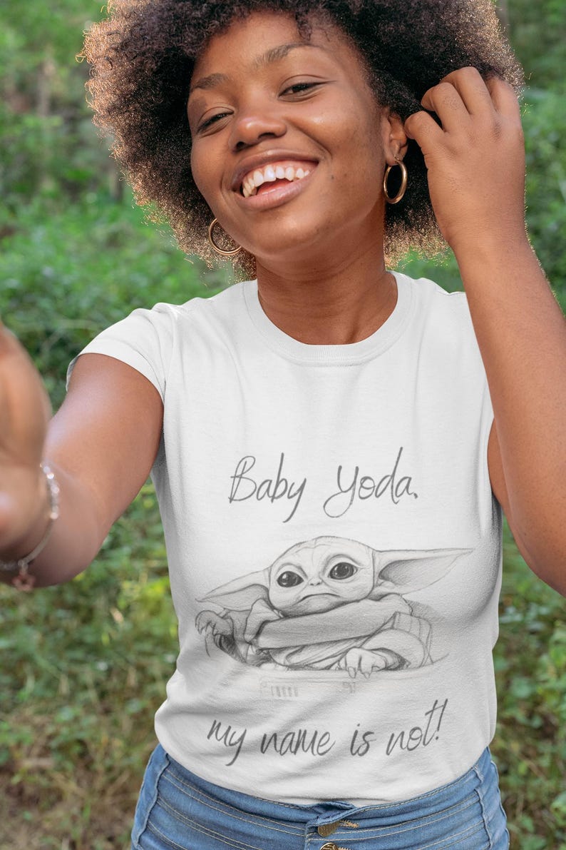 May include: White t-shirt featuring a sketch of Baby Yoda with the text "Baby Yoda, my name is not!" The shirt is worn by a person with dark curly hair and gold hoop earrings. The background is blurred green.