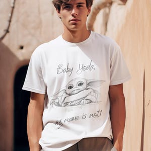 May include: White t-shirt featuring a sketch of Baby Yoda with the text "Baby Yoda, my name is not!" The shirt is a crew neck style and is made of a soft material. The shirt is worn by a person.