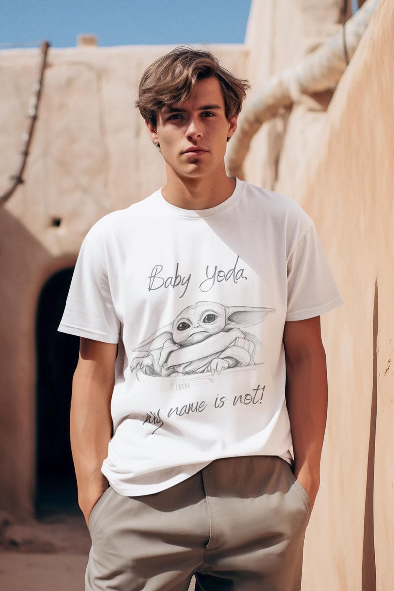 May include: White t-shirt featuring a sketch of Baby Yoda with the text "Baby Yoda, my name is not!" The shirt is a crew neck style and is made of a soft material. The shirt is worn by a person.