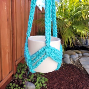 Puede incluir: Una maceta de cerámica blanca colgando de un macramé para plantas de color turquesa. El macramé está hecho de hilo grueso y retorcido.