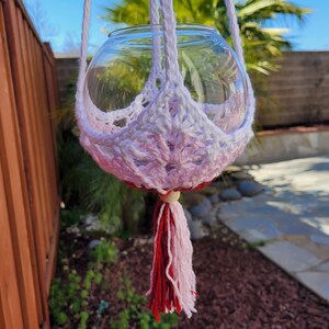 May include: A white and pink crocheted plant hanger with a glass globe and a red and white tassel.