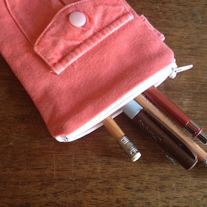 May include: A coral pink fabric pencil case with a zipper closure and a flap with a button. The case is filled with pencils and pens.