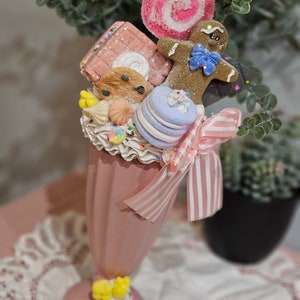Puede incluir: Un vaso de batido de leche de vidrio rosa con una cinta a rayas rosa y blanca, coronado con un hombre de jengibre, una piruleta de espiral rosa y blanca, una barra de chocolate, una galleta, un macarrón y una merengue blanca.