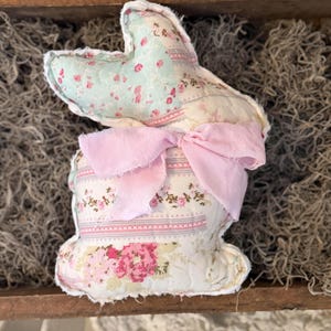 May include: A handmade fabric bunny with a patchwork design. The bunny features a light blue floral print on the ears, a pink bow, and a body with a mix of floral patterns in pink and white. The bunny is a decorative item.
