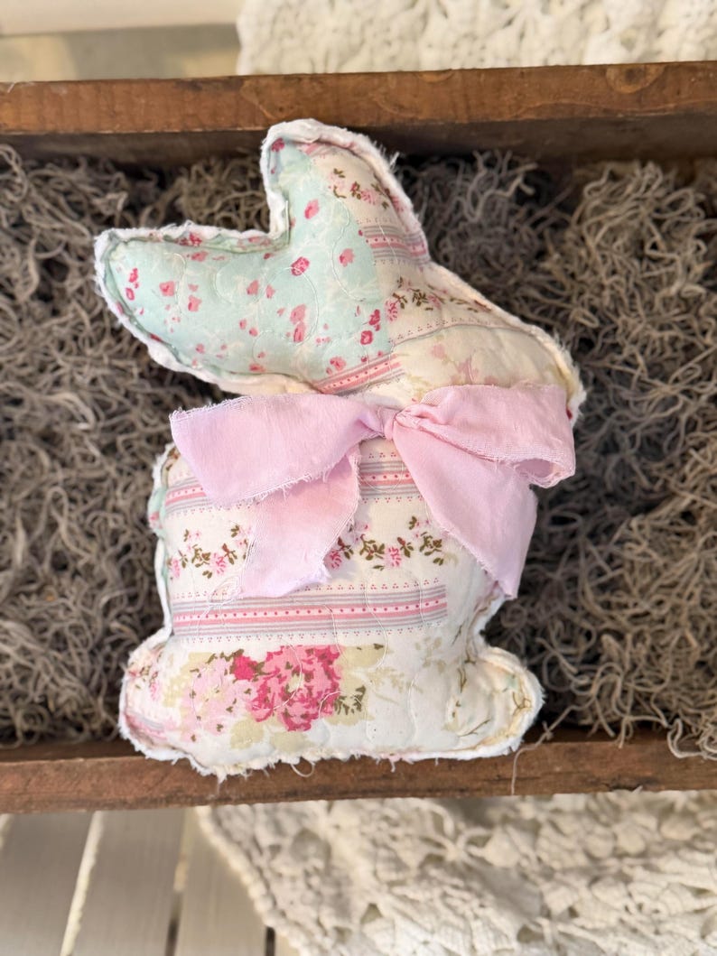May include: A handmade fabric bunny with a patchwork design. The bunny features a light blue floral print on the ears, a pink bow, and a body with a mix of floral patterns in pink and white. The bunny is a decorative item.
