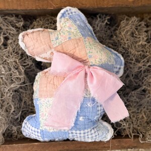May include: A handmade fabric bunny with a patchwork design in pink, blue, and floral patterns. The bunny has a large pink ribbon bow and is displayed in a wooden tray with moss.