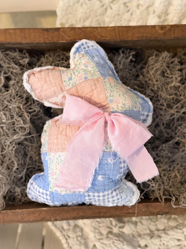 May include: A handmade fabric bunny with a patchwork design in pink, blue, and floral patterns. The bunny has a large pink ribbon bow and is displayed in a wooden tray with moss.