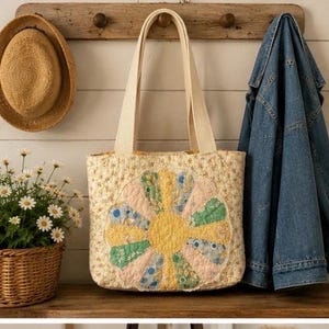 May include: A quilted tote bag featuring a cream-colored fabric with a small floral print and a large, central circular applique design composed of multi-colored, petal-shaped fabric segments in shades of yellow, green, blue, and pink.