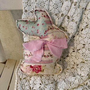 May include: A handmade fabric bunny with a patchwork design. The bunny features a light blue and floral patterned head, a pink bow, and a body with a floral pattern. The bunny is sitting on a white lace doily.