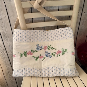 May include: A white and blue floral pattern tote bag with a beige strap. The bag features an embroidered design of blue birds and pink flowers on a white panel. The bag is sitting on a white chair.