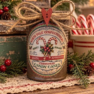 Puede incluir: Un recipiente cilíndrico marrón con una etiqueta que dice North Pole Confectionery, Christmas Candy Canes y Santa Approved. La etiqueta muestra bastones de caramelo, acebo y texto. Un lazo de cuerda de yute con un botón de corazón rojo se encuentra en la parte superior.