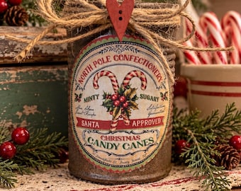 May include: A brown cylindrical container with a label reading North Pole Confectionery, Christmas Candy Canes, and Santa Approved. The label features candy canes, holly, and text. A jute twine bow with a red heart button sits on top.