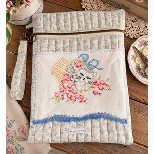 May include: A quilted pouch featuring a light blue and white floral pattern with a zippered top and a matching wrist strap. The front displays a white rectangular panel with an embroidered illustration of a kitten in a basket with pink flowers.