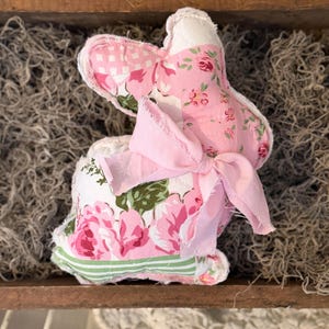 May include: A handmade fabric bunny with pink floral patterns and a pink bow. The bunny is made of various fabrics, including a pink and white gingham, and a floral print with pink roses and green leaves. The bunny is sitting in a wooden tray.