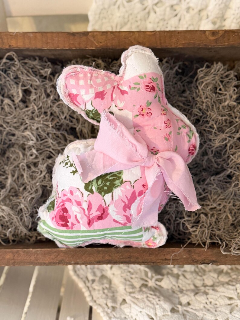 May include: A handmade fabric bunny with pink floral patterns and a pink bow. The bunny is made of various fabrics, including a pink and white gingham, and a floral print with pink roses and green leaves. The bunny is sitting in a wooden tray.