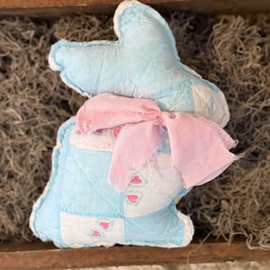 May include: A light blue quilted bunny-shaped plush toy with a pink bow. The bunny has a white trim and a patchwork design with pink heart details. The toy is placed in a wooden tray with a moss-like material.