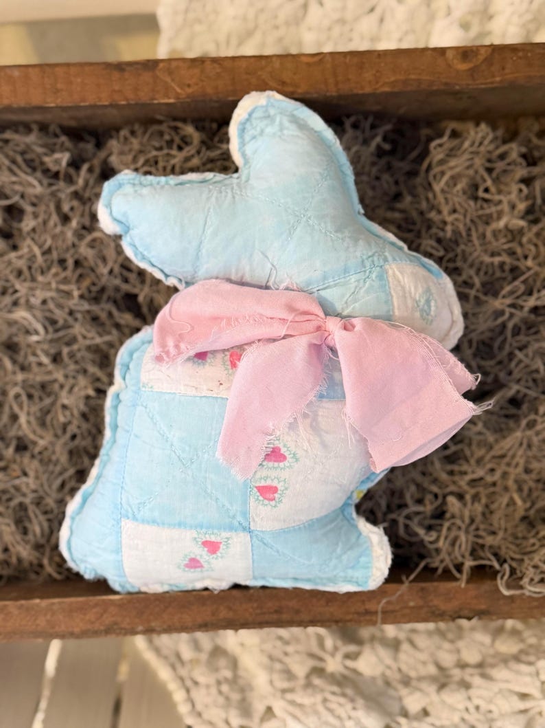 May include: A light blue quilted bunny-shaped plush toy with a pink bow. The bunny has a white trim and a patchwork design with pink heart details. The toy is placed in a wooden tray with a moss-like material.