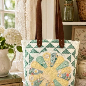 Puede incluir: Un bolso de tela acolchado con un gran dise&ntilde;o central de apliques florales en amarillo y verde. El bolso est&aacute; confeccionado con tela blanca y patrones geom&eacute;tricos triangulares verdes. Incluye dos correas de cuero marr&oacute;n con remaches.