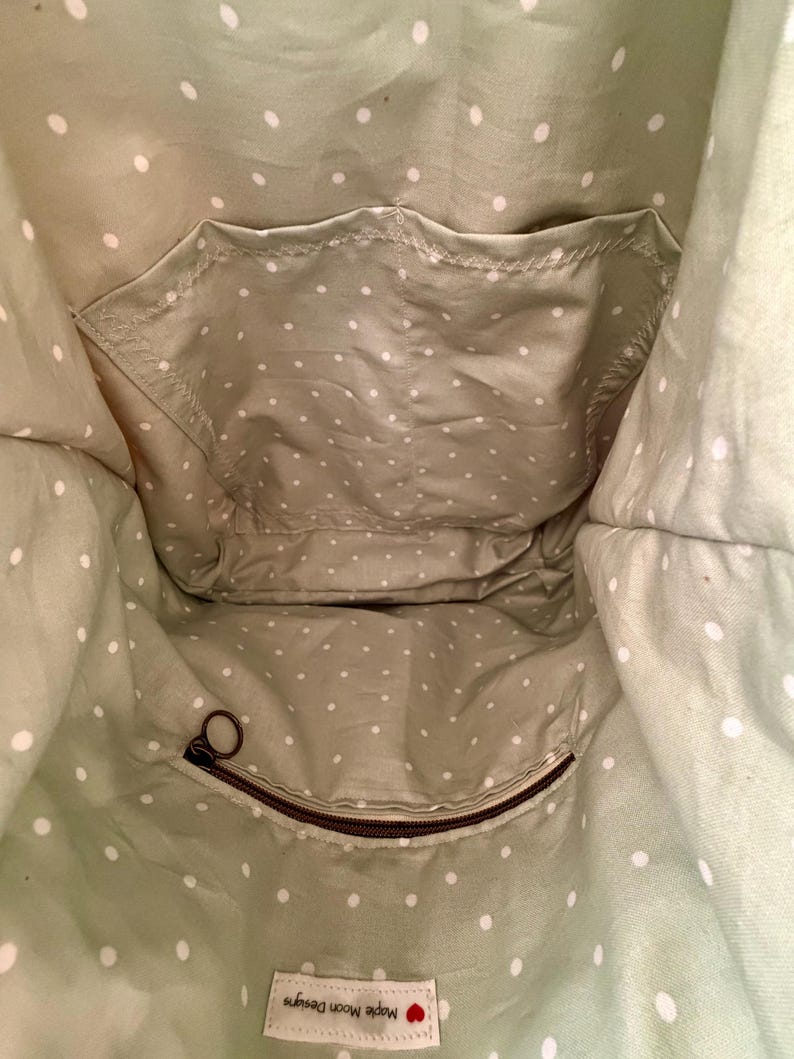 Puede incluir: El interior de un bolso de tela verde salvia con un patr&oacute;n de lunares blancos. Incluye un bolsillo abierto, un bolsillo con cremallera con un tirador marr&oacute;n y una etiqueta de tela blanca rectangular que dice Maple Moon Designs.