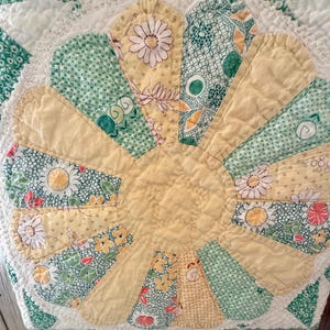 Puede incluir: Un textil acolchado que presenta un motivo floral central con p&eacute;talos alternos en amarillo s&oacute;lido y varios patrones florales en verde y blanco. La colcha tiene un borde festoneado y est&aacute; cosida con un acabado acolchado texturizado.