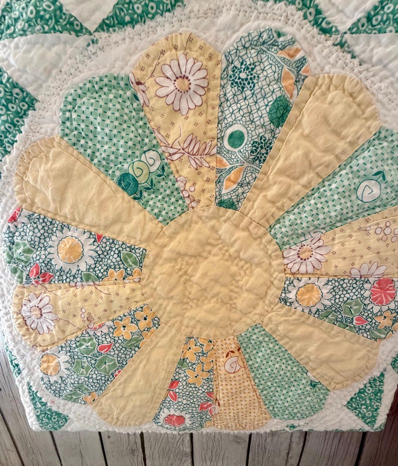 Puede incluir: Un textil acolchado que presenta un motivo floral central con p&eacute;talos alternos en amarillo s&oacute;lido y varios patrones florales en verde y blanco. La colcha tiene un borde festoneado y est&aacute; cosida con un acabado acolchado texturizado.