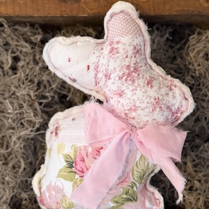 May include: A handmade fabric bunny with a pink floral pattern and a pink bow. The bunny is made of soft, quilted fabric and has a whimsical, vintage aesthetic. The bunny is a decorative item.