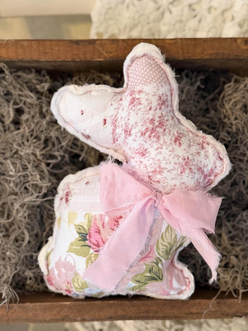 May include: A handmade fabric bunny with a pink floral pattern and a pink bow. The bunny is made of soft, quilted fabric and has a whimsical, vintage aesthetic. The bunny is a decorative item.
